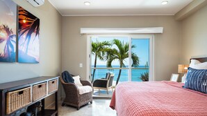 A bedroom with a large bed, a chair, a small table, and a view of the ocean.
