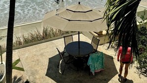 A beach view with a table and chairs under a large umbrella, palm trees, and the ocean.