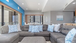 A modern living room with a grey sectional sofa, blue patterned cushions, a dining area with a table and chairs, and a view of a high-rise building through a glass door. - Las Palomas Phase II Cortez 507 Exceptional Oceanview 3BR 3BA Unit (Puerto Peñasco)