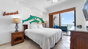 Une chambre d’hôtel avec un lit, deux tables de chevet, un bureau, une grande fenêtre donnant sur la mer et un mur orné d’un tableau représentant un dauphin.