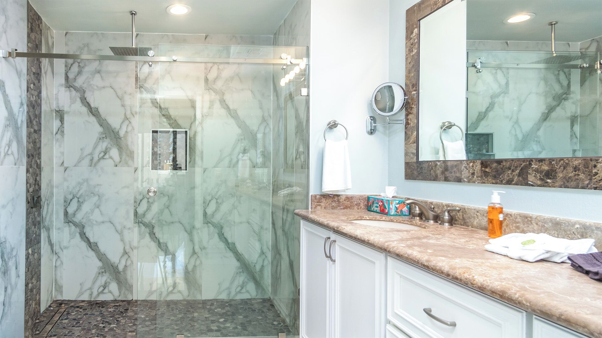 Une salle de bain moderne dotée d’une douche à parois de verre, de murs en marbre, d’un meuble-lavabo avec un plan de travail en marbre et d’un grand miroir.