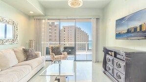 Een moderne woonkamer met een bank, een glazen salontafel en een donker houten kast. Een groot raam biedt uitzicht op hoge gebouwen en een strand.