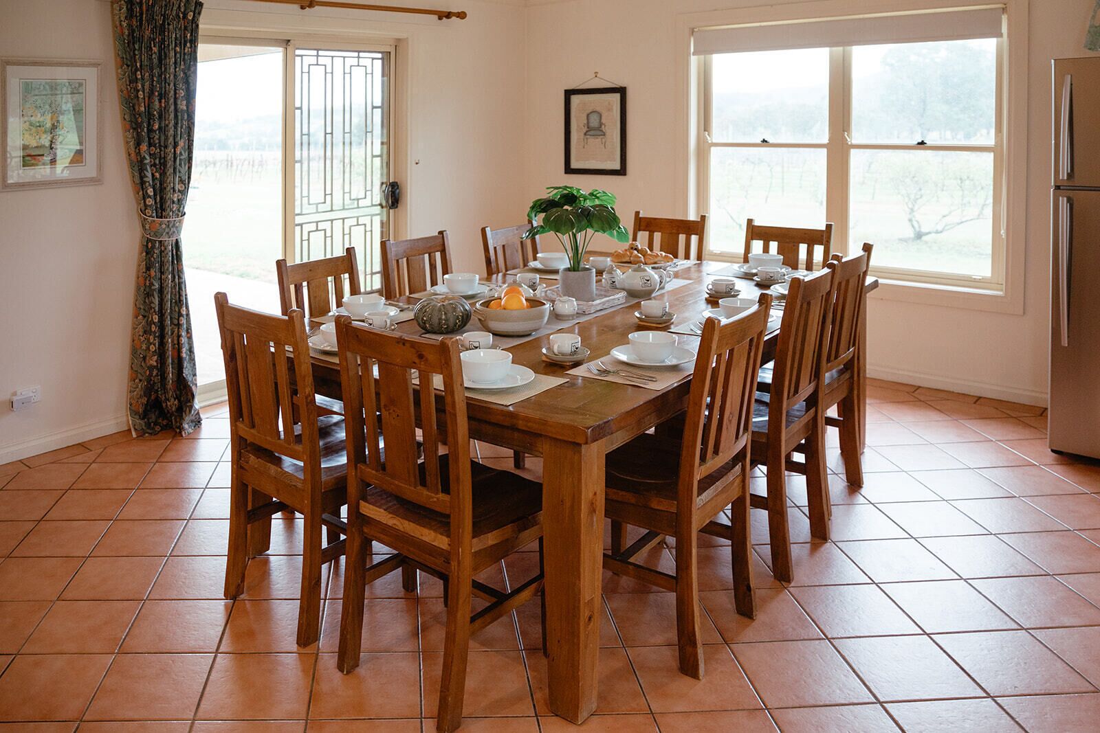 Ein Esszimmer mit einem Holztisch, gedeckt für sechs Personen, umgeben von Holzstühlen. Auf dem Tisch steht ein Blumentopf, und der Raum verfügt über große Fenster mit Vorhängen.