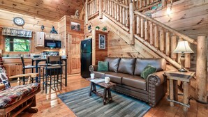 A cozy cabin interior with a wooden staircase, a dining area, a living room with a leather sofa, and a kitchen with wooden cabinets and appliances. - Family friendly cabin in great location! Arcade~Hot Tub~Foosball~Fireplace (Sevierville)