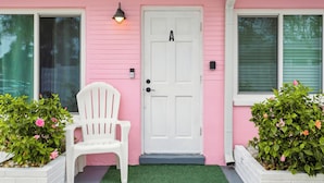 Uma casa rosa com porta branca marcada 'A', uma cadeira branca e dois floreiros com flores cor-de-rosa.