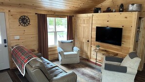 A cabin interior with a sofa, two armchairs, a television, a clock, and a window with curtains. - Miners Cabin - Pictured Rocks Lodging (Munising)