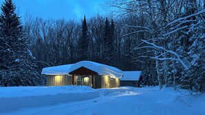 A snow-covered cabin with a wooden exterior and a sloped roof, surrounded by a snowy landscape and dense forest. - Miners Cabin - Pictured Rocks Lodging (Munising)