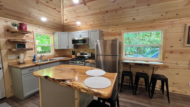 A wooden cabin kitchen with a central island, stainless steel appliances, and a window overlooking greenery. - Miners Cabin - Pictured Rocks Lodging (Munising)