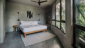 Ein modernes Schlafzimmer mit einem großen Bett, einem Deckenventilator und Blick auf Grün hinter großen Fenstern.