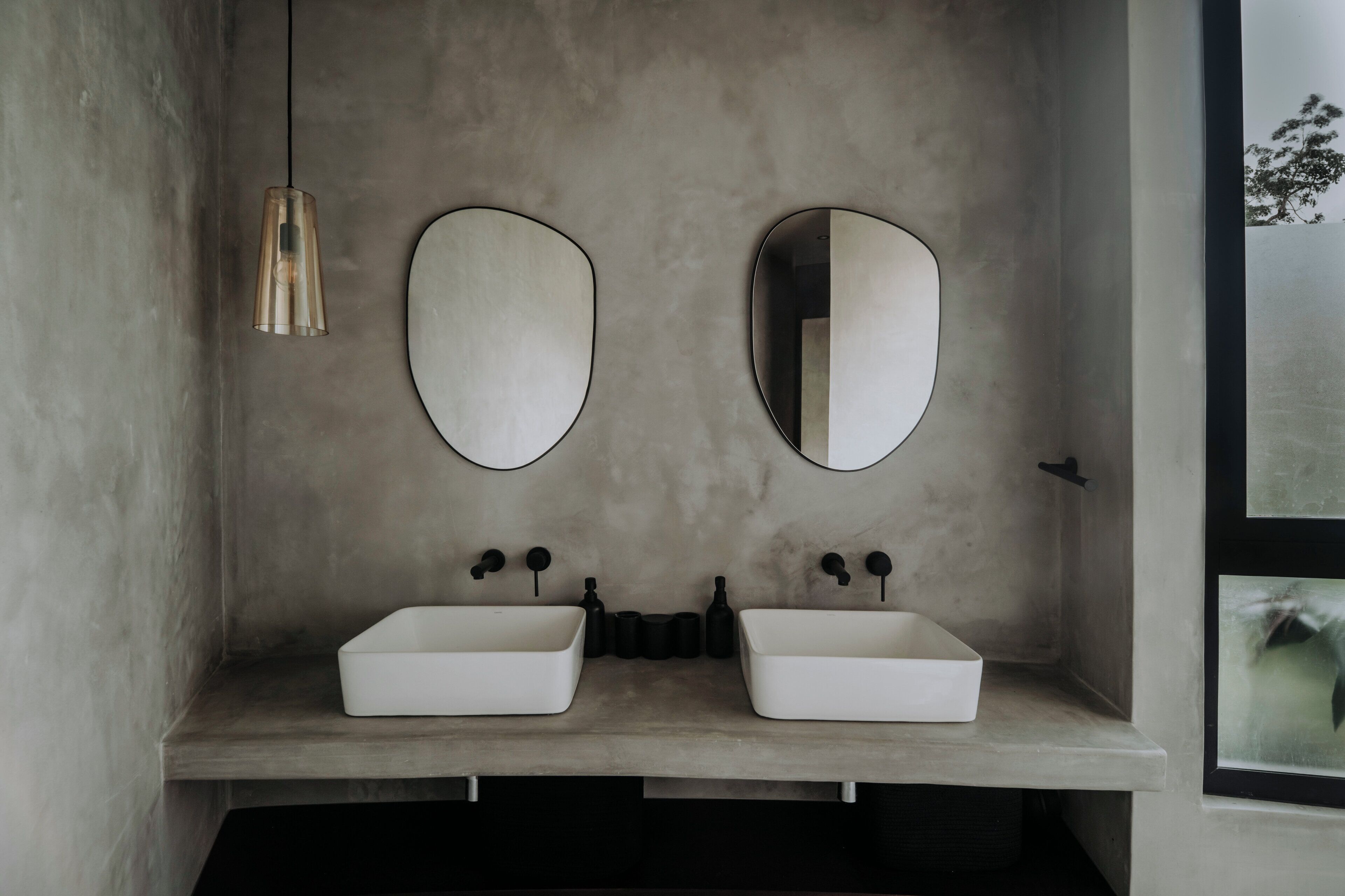 A bathroom with two white sinks, a single hanging light, and two round mirrors on a concrete wall.