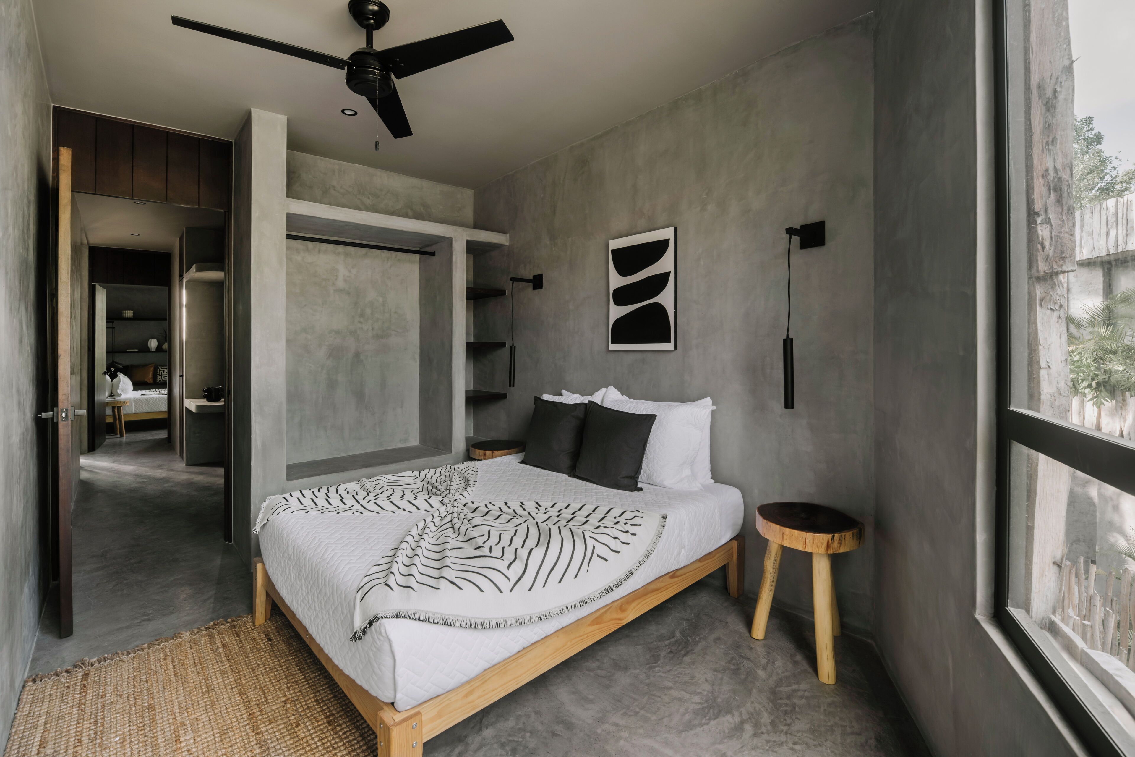 A modern bedroom with a large bed, a ceiling fan, a view through a window, and a wooden stool.
