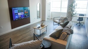A modern living room with a flat-screen TV mounted on the wall, a sofa set, a coffee table, and a large window providing natural light. - StayAE - High Rise (Baton Rouge)