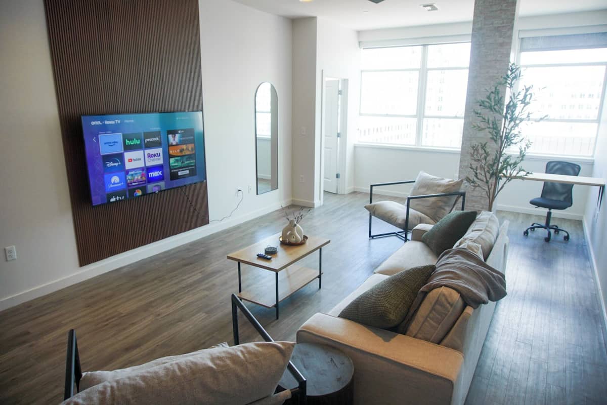 A modern living room with a flat-screen TV mounted on the wall, a sofa set, a coffee table, and a large window providing natural light.