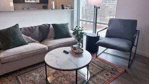 A modern living room with a sofa, armchair, coffee table, and a view of the city through a large window.