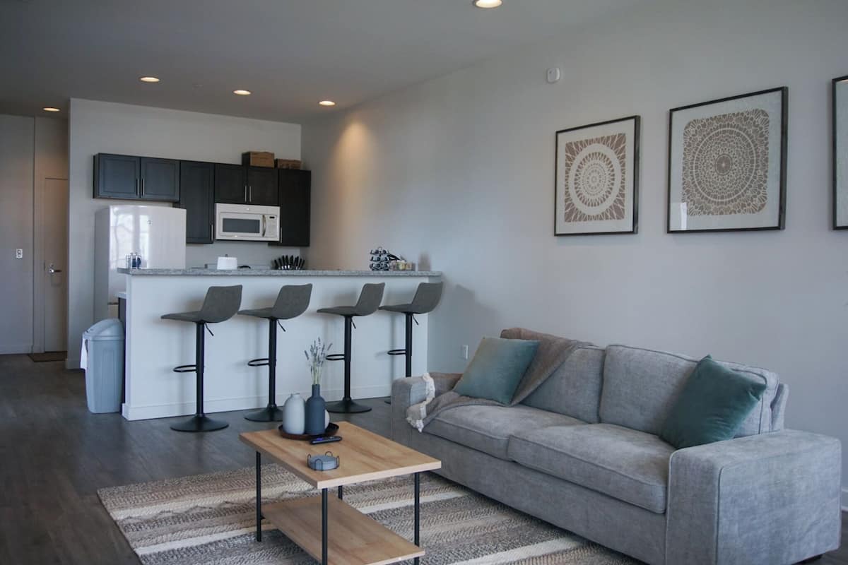 A modern apartment with a kitchen, a grey sofa, and two bar stools.