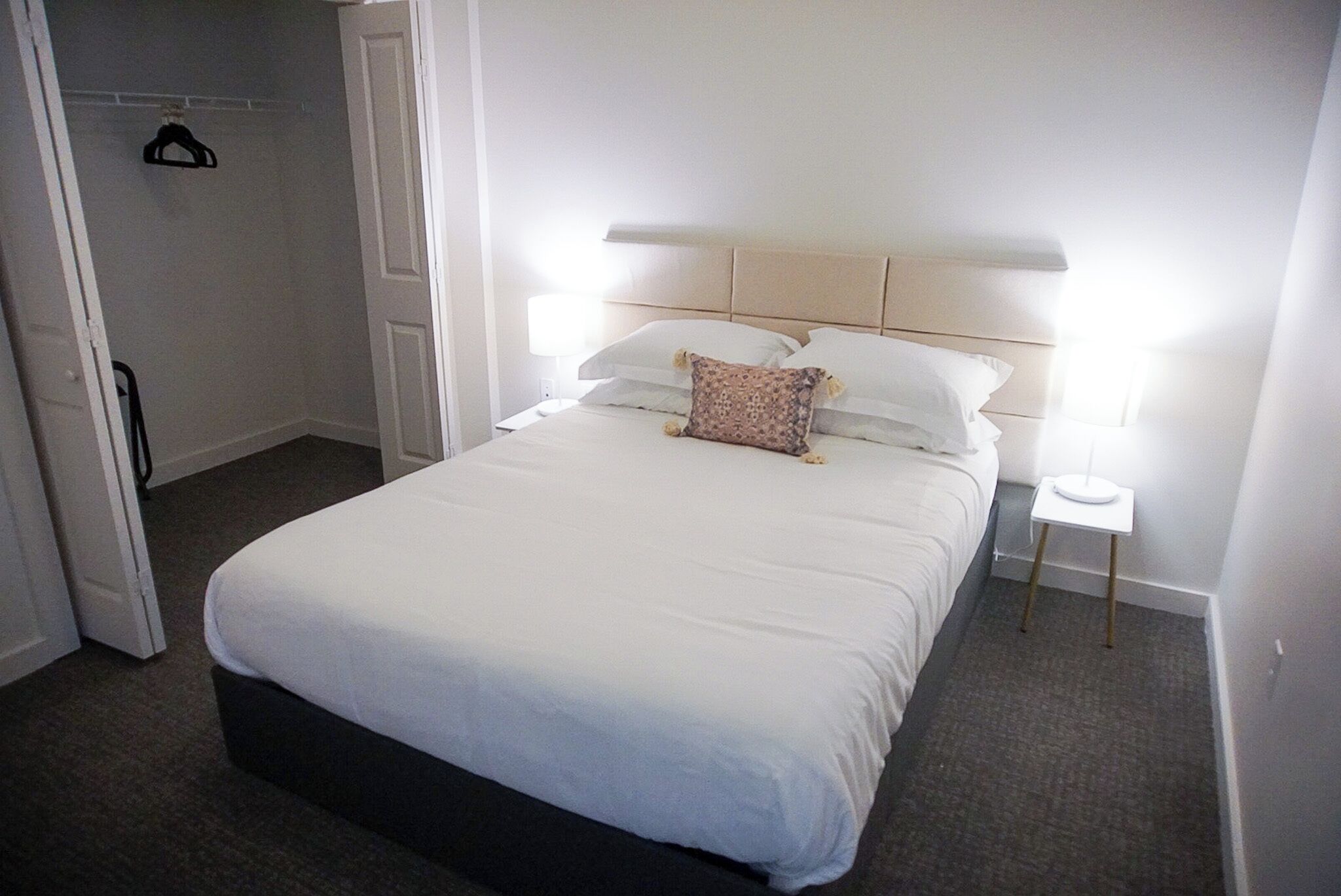 A neatly made bed with white linens and a single decorative pillow, a bedside table with a lamp, and a door leading to a closet with a hanger.