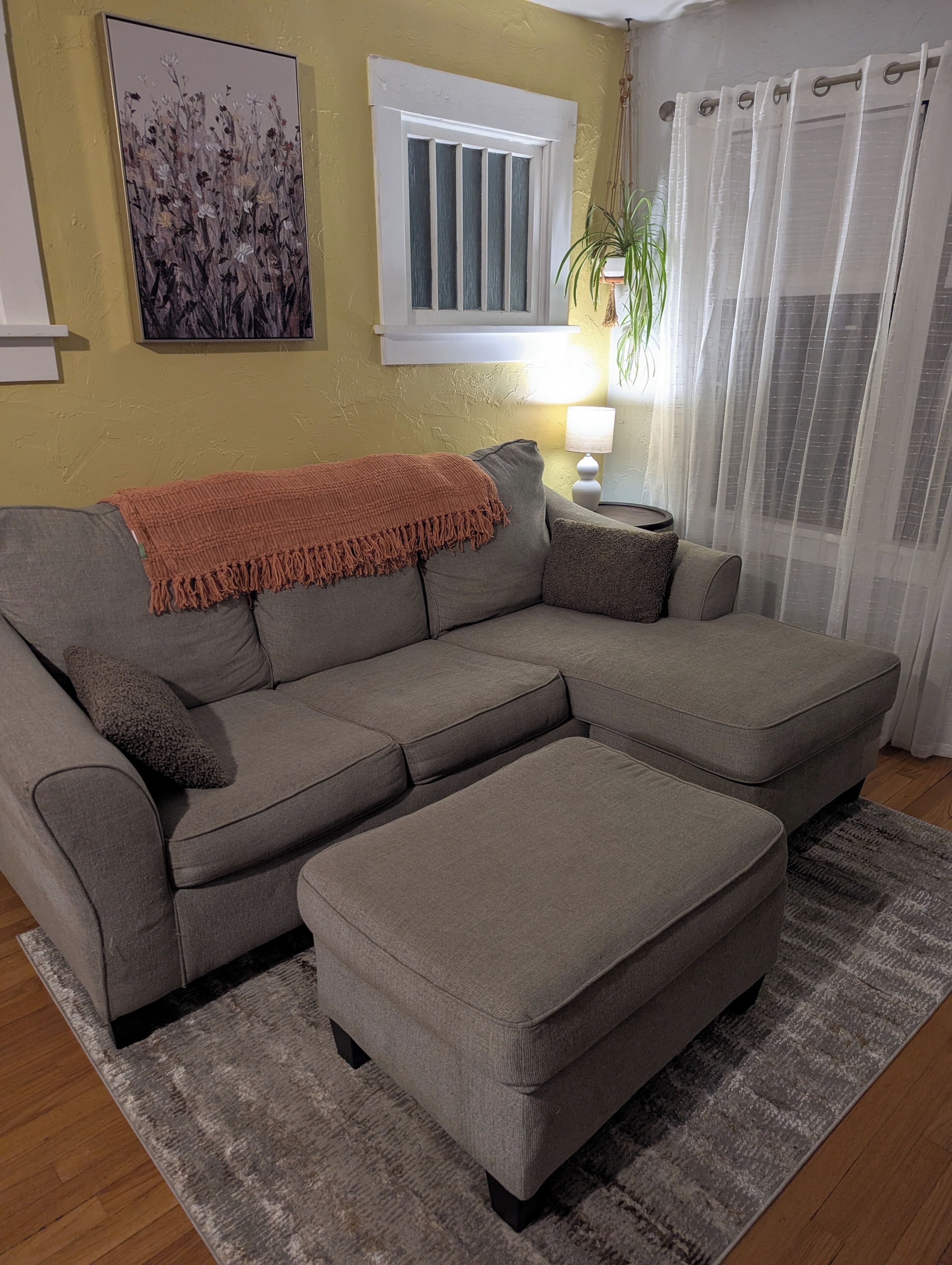 Una sala de estar con un sofá modular, un pequeño reposapiés, una lámpara, una planta y una ventana con cortinas.