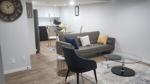 Una sala moderna con un sofá gris, un sillón negro y una mesa de centro de vidrio. En el fondo, hay una zona de comedor con una mesa y sillas.