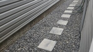 Un sendero estrecho con una pared de metal a un lado y un suelo de grava con piedras para pisar.