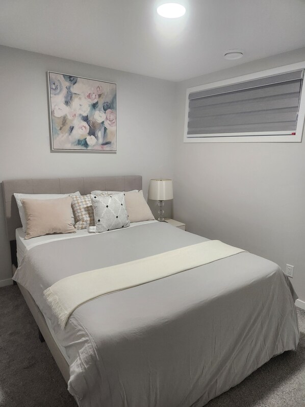 Un dormitorio con cama, mesita de noche, lámpara, ventana con persianas y un cuadro en la pared.