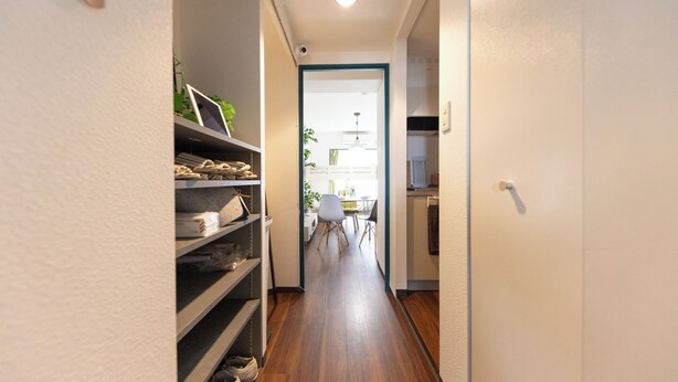 Un estrecho pasillo con suelo de madera, estanterías a la izquierda y una puerta que da a una habitación con una mesa y sillas.