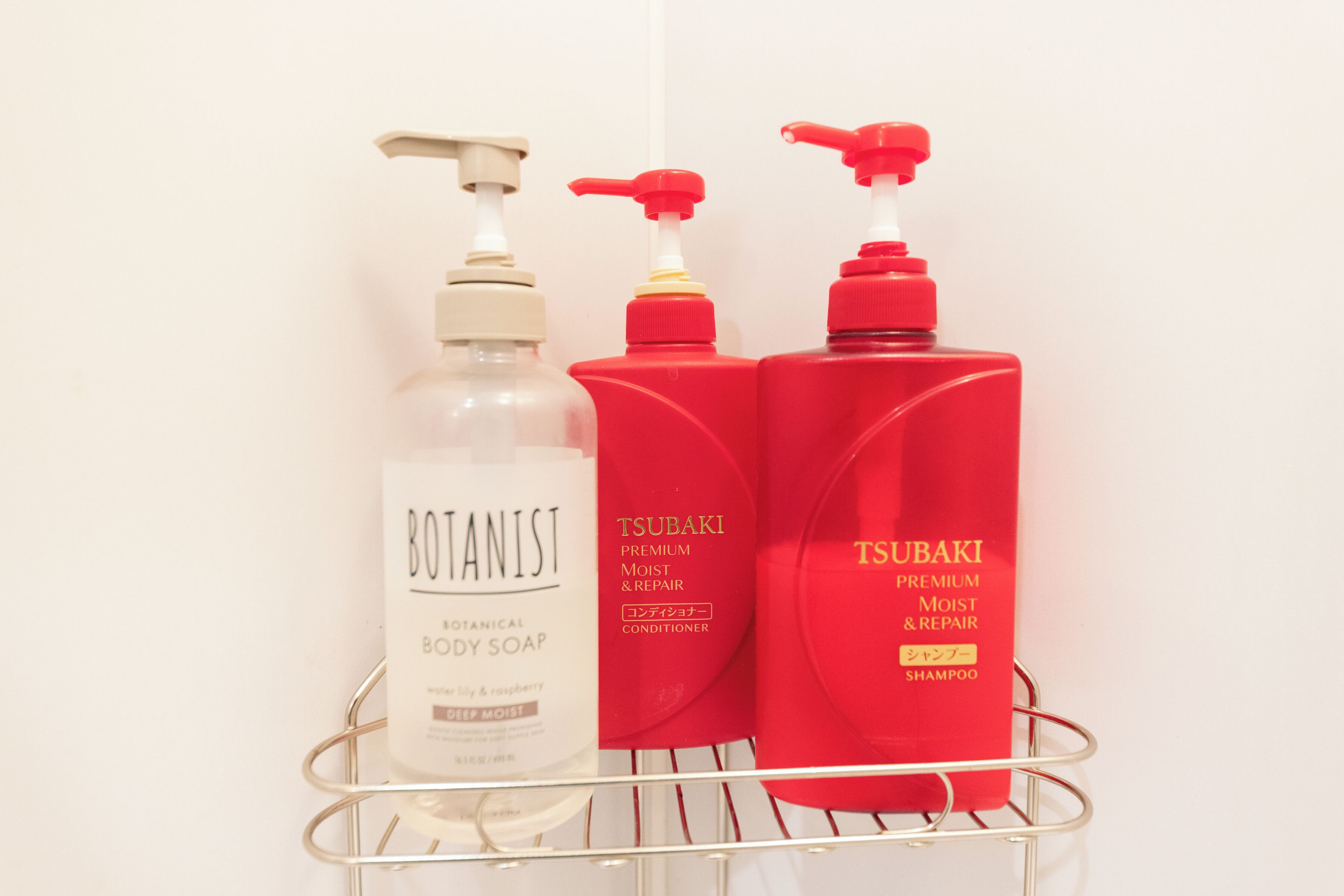 Un estante de baño con tres botellas de productos para el cuidado corporal: jabón de baño Botanist, acondicionador Tsubaki premium hidratación y reparación, y champú Tsubaki premium hidratación y reparación.
