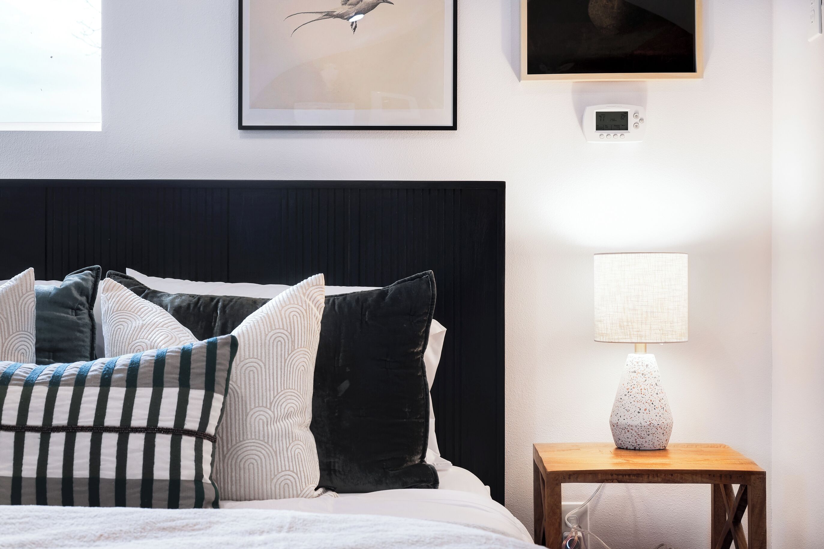 Ein ordentlich bezogenes Bett mit verschiedenen Kissen, ein Nachttisch mit Lampe und ein gerahmtes Bild an der Wand.