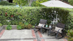 A patio with two wicker chairs and a beige umbrella, surrounded by lush greenery and a brick border. - Santa Barbara Mission Studio (Santa Barbara)