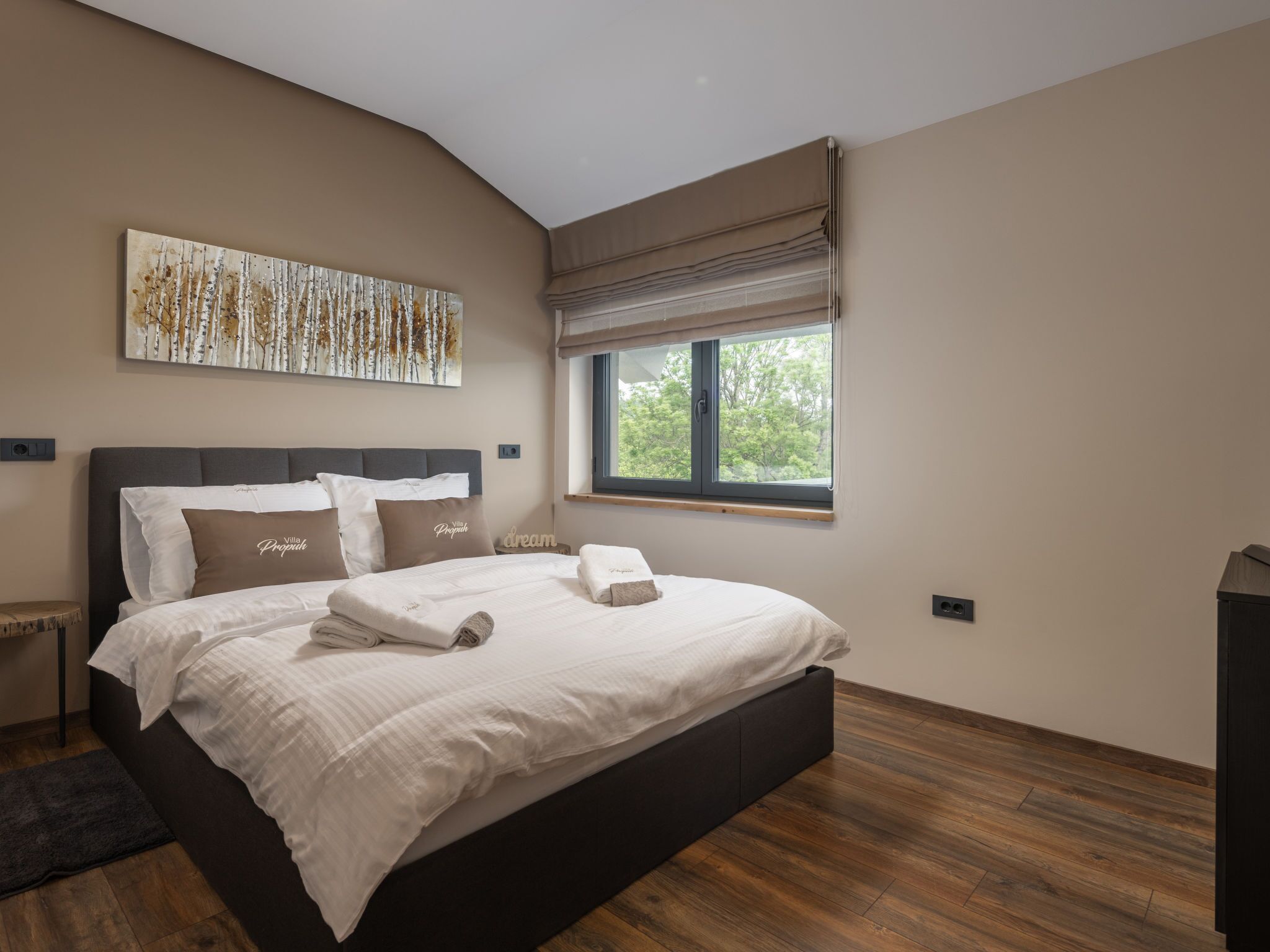 A modern bedroom with a large bed, a window with blinds, a wooden floor, and a wall-mounted artwork.