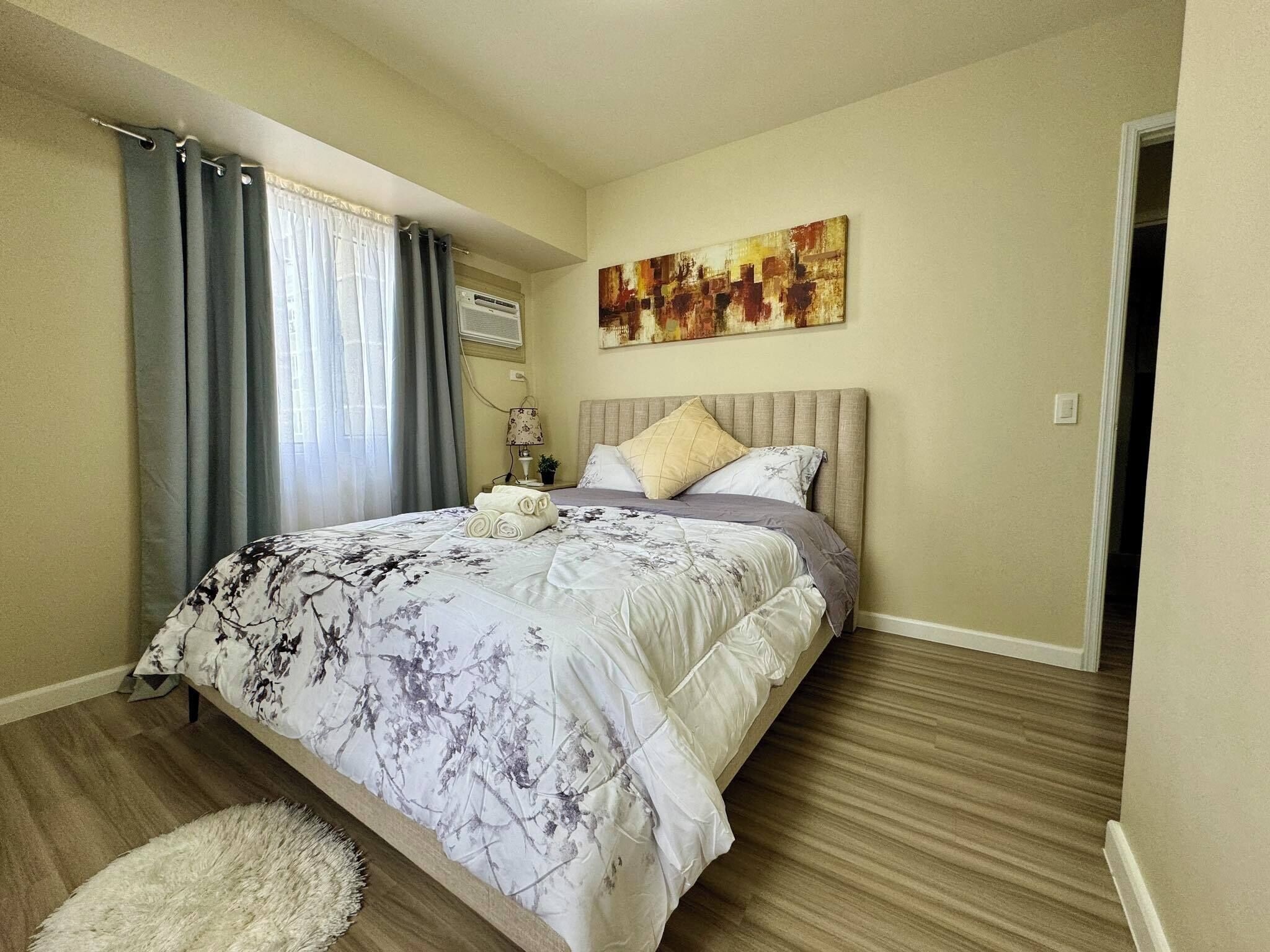 A bedroom with a bed, a painting on the wall, and a window with curtains.