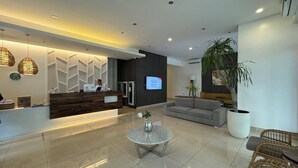 A modern reception area with a wooden reception desk, a grey sofa, a round glass table, and a large potted plant. - Condotel Avida Riala Tower 4-1103 (Cebu City)