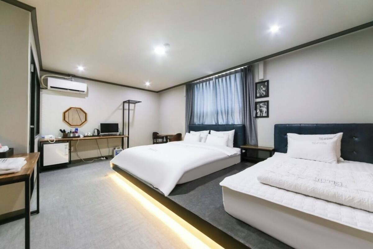 A hotel room with two beds, a desk with a computer, an air conditioning unit, and a window with curtains.