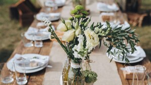 Una mesa de madera dispuesta para una comida, con platos blancos, vasos y un centro de mesa con flores en frascos de vidrio.