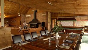 Un comedor con una larga mesa de madera dispuesta para una comida, una chimenea de ladrillo y vistas de un jardín a través de una ventana con celosía.