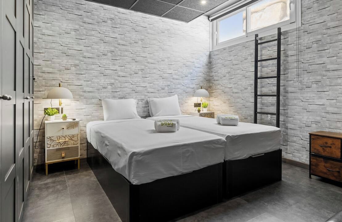Une chambre avec un mur en briques, deux lits, des lampes de chevet, une échelle et une commode en bois.