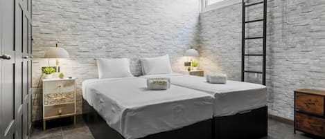 Une chambre avec un mur en briques, deux lits, des lampes de chevet, une échelle et une commode en bois.