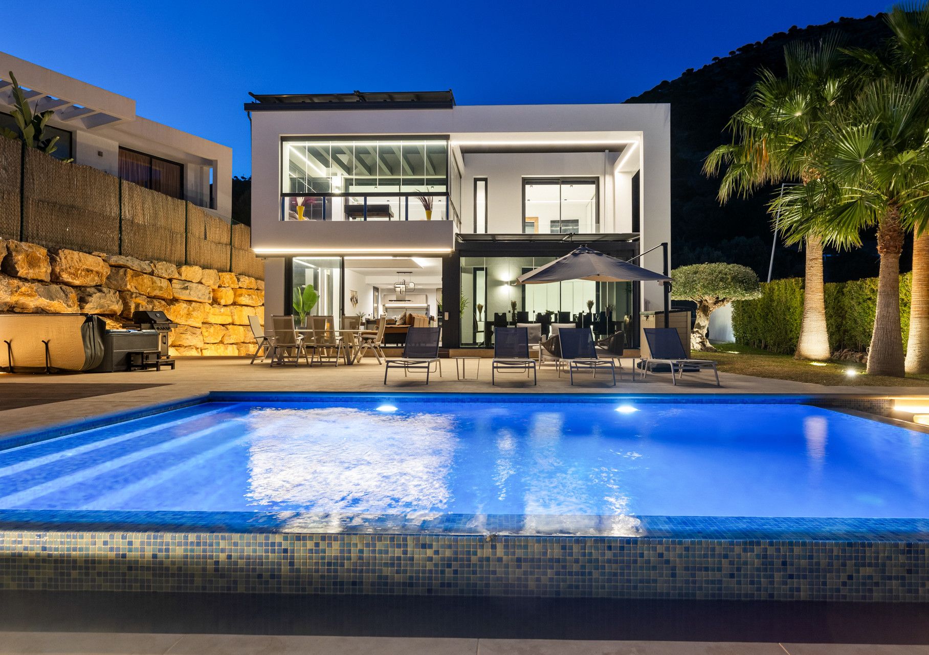 A modern two-story house with a swimming pool, outdoor seating, and a stone wall.