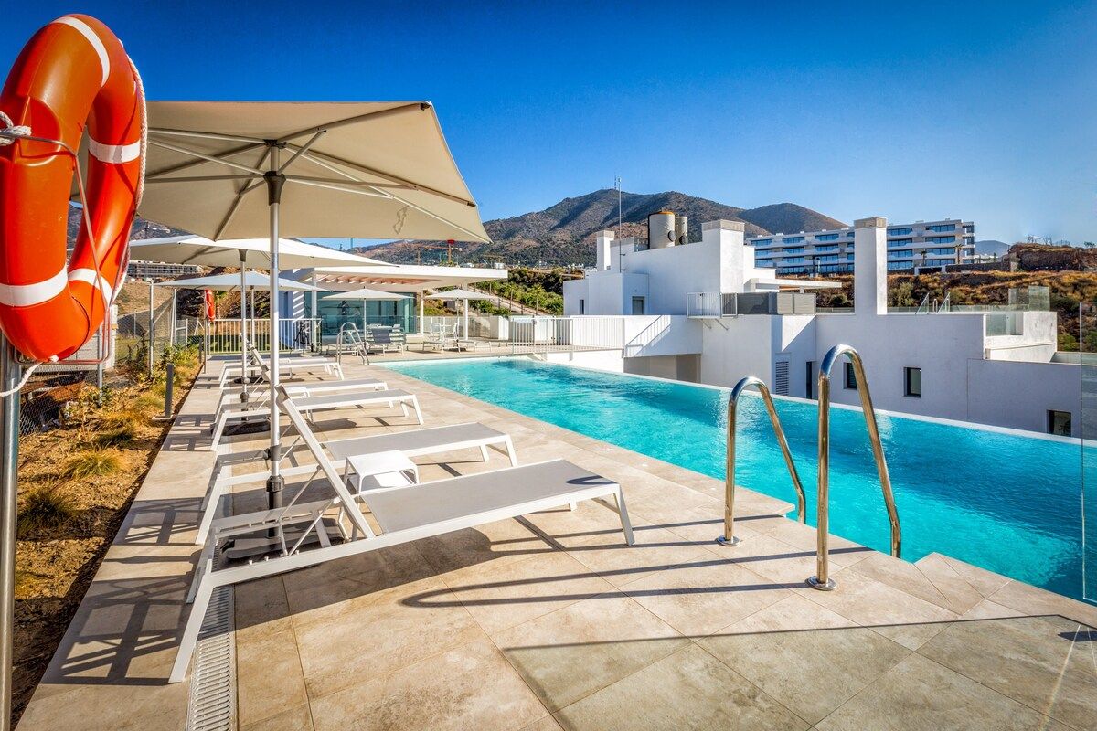 A swimming pool with sun loungers and a lifebuoy, surrounded by a white building with a mountain view in the background.