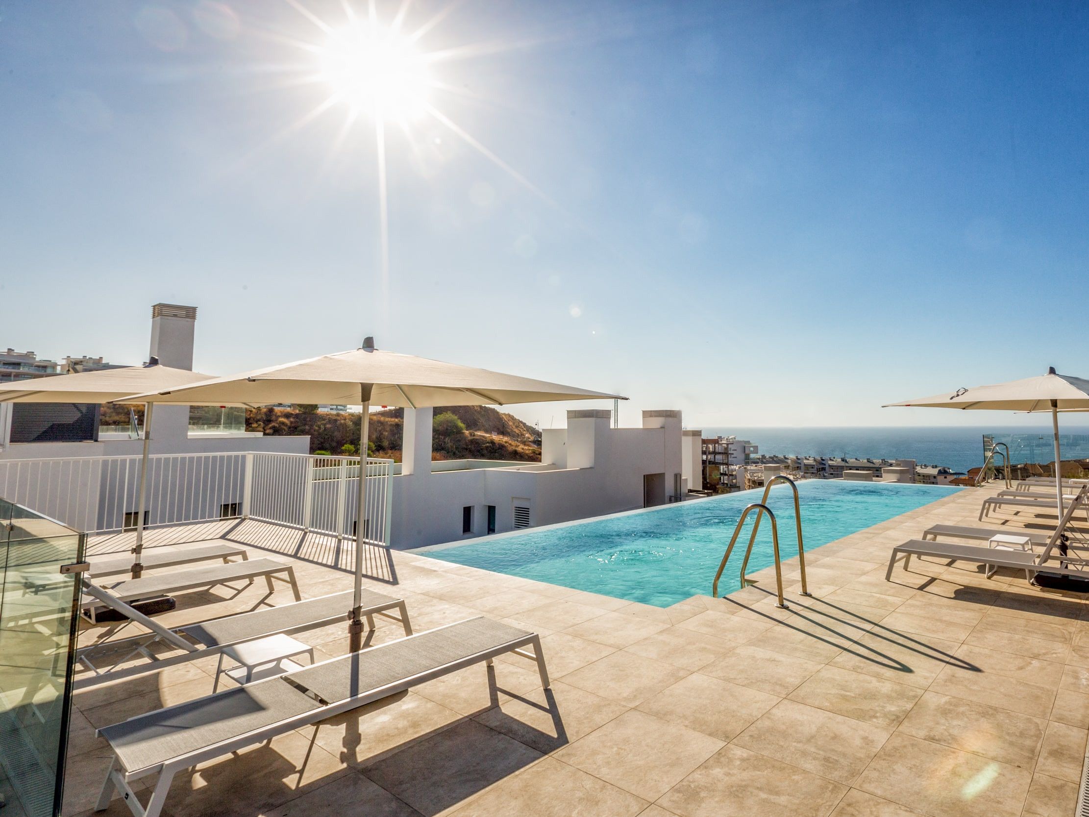 Uma área de piscina no terraço, com espreguiçadeiras e guarda-sóis, vista clara do oceano e piso revestido.