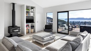 Ein modernes Wohnzimmer mit einem schwarzen Holzofen, grauen Sofas, einem Flachbildfernseher und einer großen Schiebeglastür, die auf einen Balkon mit Stadtblick führt.