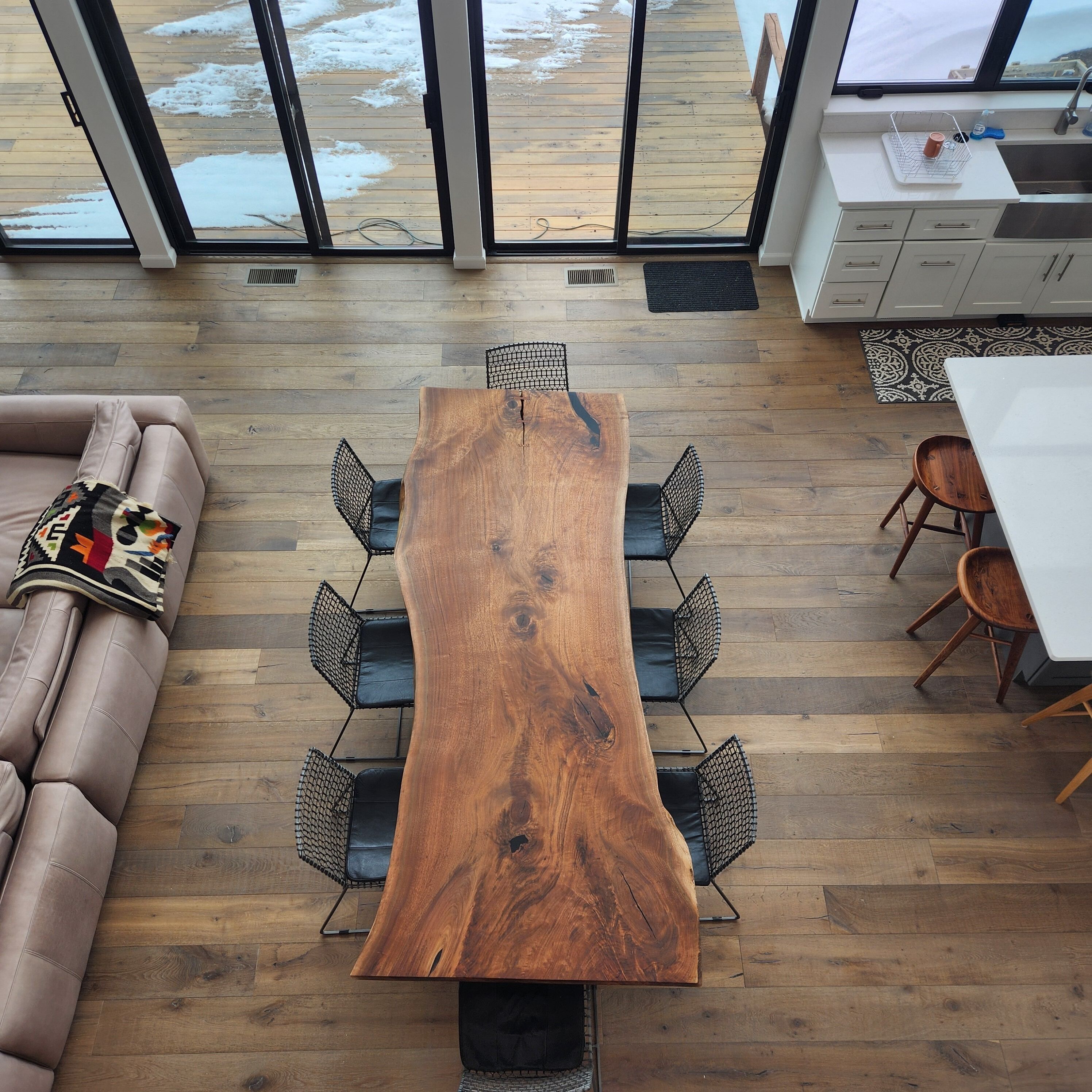 A modern interior with a large wooden dining table, cushioned seating, and a view of a wooden deck through large glass doors.