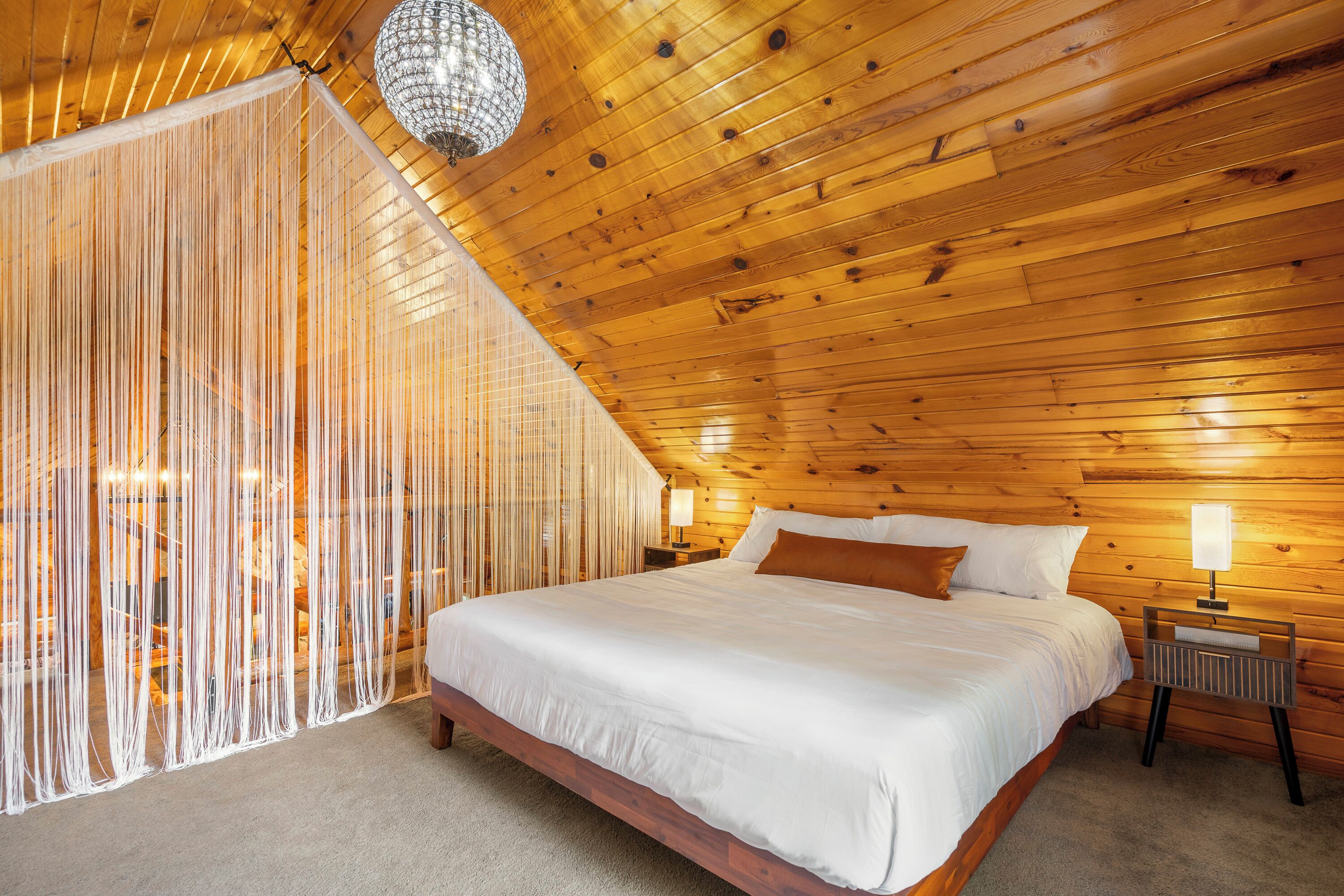 A bedroom with a wooden ceiling, a bed with white bedding, a nightstand with a lamp, and a decorative hanging light fixture.