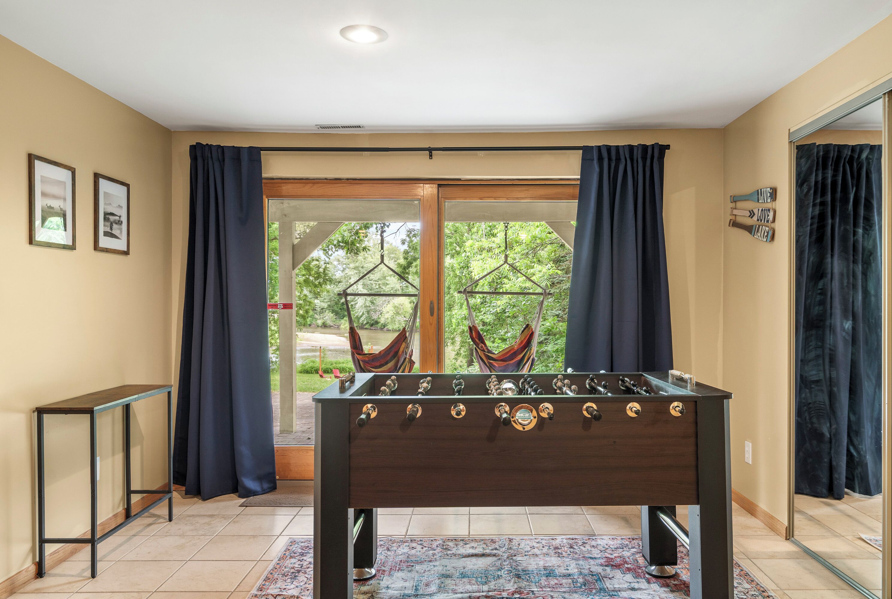 Une pièce avec une table de baby-foot, une petite table d’appoint et une vue sur un hamac extérieur, visible à travers une porte vitrée.