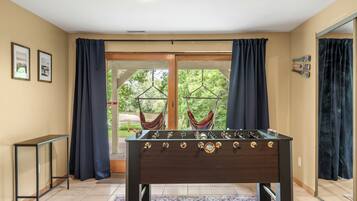 Une pièce avec une table de baby-foot, une petite table d’appoint et une vue sur un hamac extérieur, visible à travers une porte vitrée.