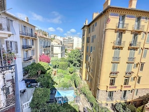 A narrow urban street with multi-story buildings, balconies, and greenery.