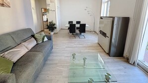 En moderne stue med sofa, sofabord i glas, et spiseområde med stole og et køkken med køleskab.