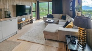 Una sala de estar moderna con una televisión de pantalla plana, un sofá, una mesa de centro y un ventanal con vistas a un área boscosa.