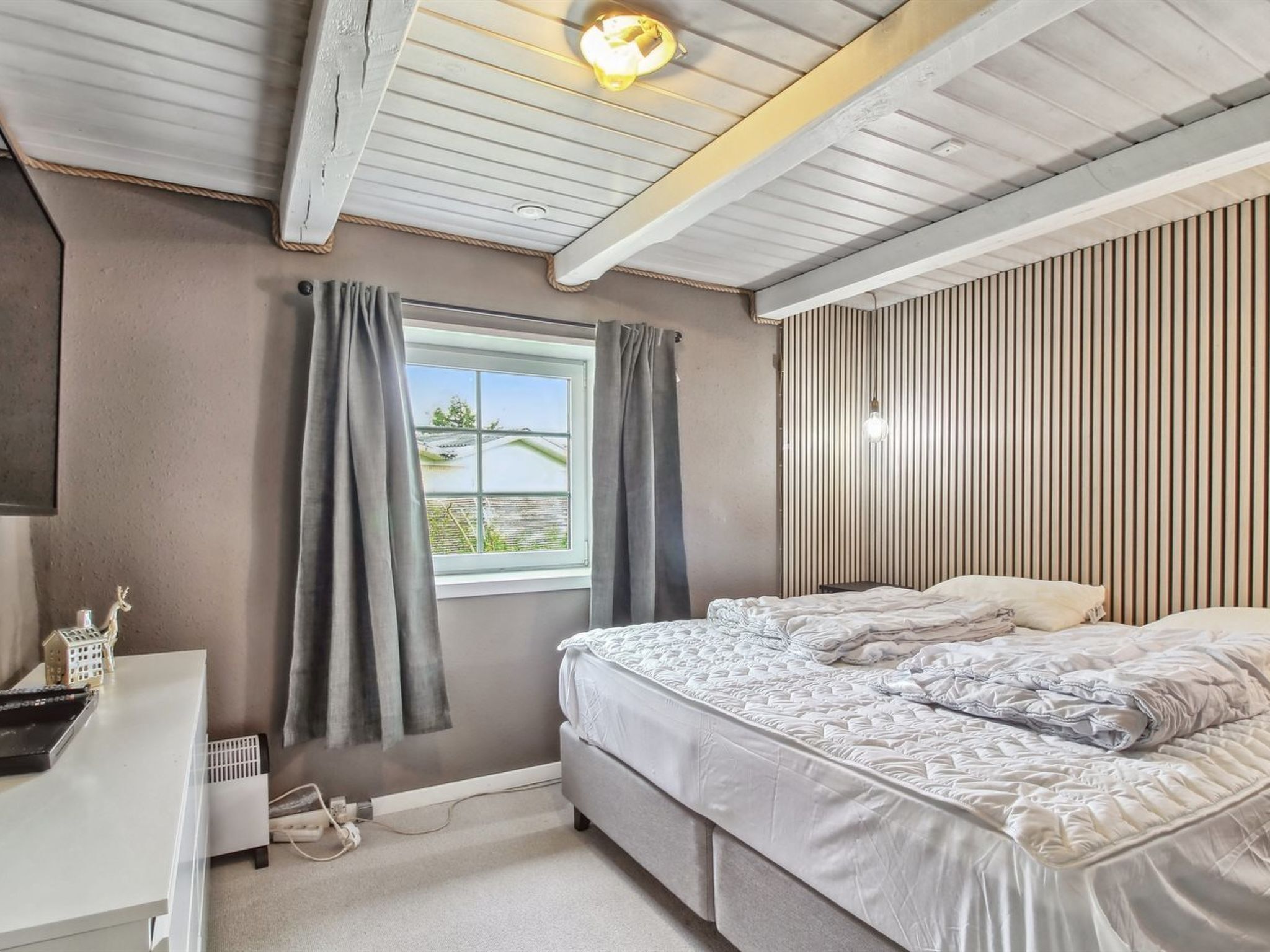 Un dormitorio con cama, escritorio, ventana con cortinas y techo inclinado con vigas a la vista.