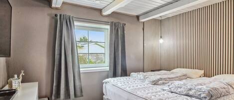 Un dormitorio con cama, escritorio, ventana con cortinas y techo inclinado con vigas a la vista.