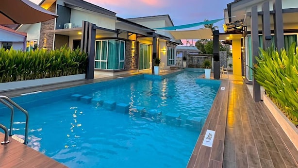 A swimming pool with a wooden deck, surrounded by modern buildings with large windows and a sunset sky. - Tijami Villa & Resort (Setiu)
